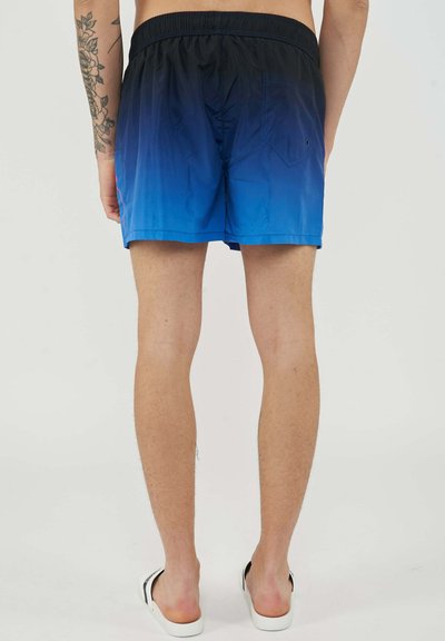 Man standing wearing blue ombre swim shorts and white slide sandals, showing tattooed left arm and bare legs.