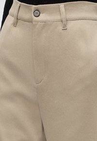Taille de pantalon beige avec fermeture par bouton, passants de ceinture et coutures visibles sur un fond de haut noir.