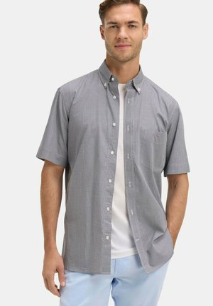 Homme portant une chemise à manches courtes à carreaux noir et blanc boutonnée sur un t-shirt blanc et un pantalon bleu clair, debout devant un fond uni.