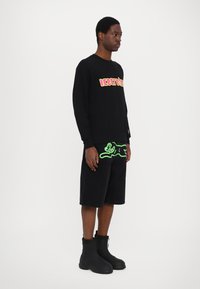 Svart genser med livlig rosa og gul "iskrem" tekst, parret med svarte shorts med et grønt grafisk design. Brukt med svarte støvler.
