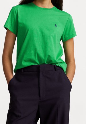 Kvinde iført en knaldgrøn kortærmet t-shirt med et lille blåt logo på brystet, stoppet ned i højtaljede sorte bukser med hænderne i lommerne.