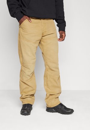 Beiget työhousut kestävästä kankaasta, joissa on rento istuvuus, vahvistetut saumat ja etutaskut, yhdistettynä mustiin jalkineisiin.