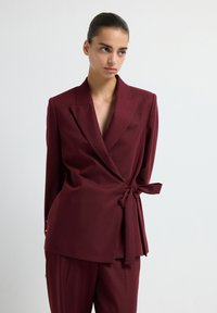Blazer avvolgente bordeaux in un tessuto morbido con maniche lunghe, scollatura a V profonda e dettaglio con fiocco laterale. Abbinato a pantaloni coordinati.
