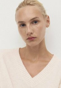 Weicher rosa Wollpullover mit V-Ausschnitt und gerippten Akzenten. Das Model hat helle Haut und hellblondes Haar, vor einem neutralen Hintergrund.