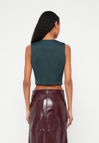 Frau mit langen lockigen Haaren, die ein ärmelloses, dunkelgrünes, bauchfreies Oberteil und einen hoch taillierten, glänzenden burgunderfarbenen Rock trägt, von hinten gezeigt.