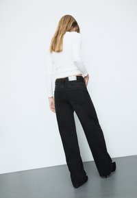 Jeans neri a gamba larga con vita alta e design a cinque tasche. Sul retro presentano due tasche applicate e un'etichetta in vita. Abbinati a una maglietta bianca a maniche lunghe.