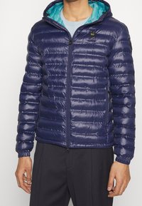 Giacca puffer blu navy con cappuccio, dotata di una fodera turchese a contrasto, chiusura frontale con zip e polsini elasticizzati. Texture liscia e lucida.