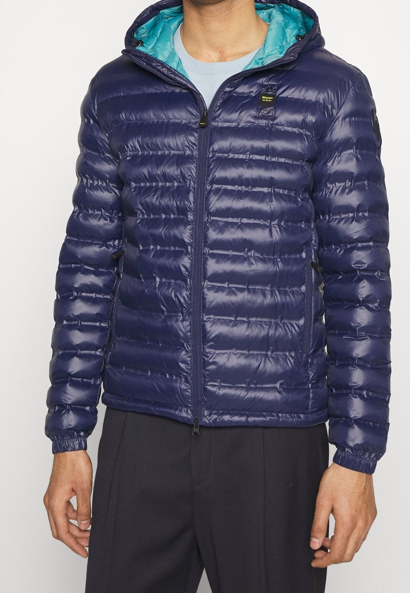 Giacca puffer blu navy con cappuccio, dotata di una fodera turchese a contrasto, chiusura frontale con zip e polsini elasticizzati. Texture liscia e lucida.