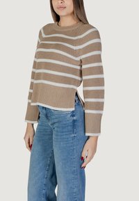 Maglione a strisce beige con linee bianche, texture a coste, scollo rotondo e design leggermente corto con fessure laterali. Indossato con jeans blu.