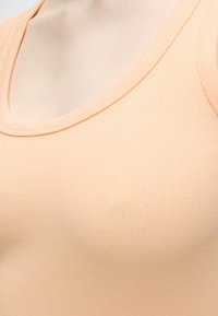 Κοντινό πλάνο στο πάνω μέρος του στήθους και του λαιμού ενός ατόμου που φοράει ένα ανοιχτό ροδακινί ριπ τοπ με στρογγυλή λαιμόκοψη.