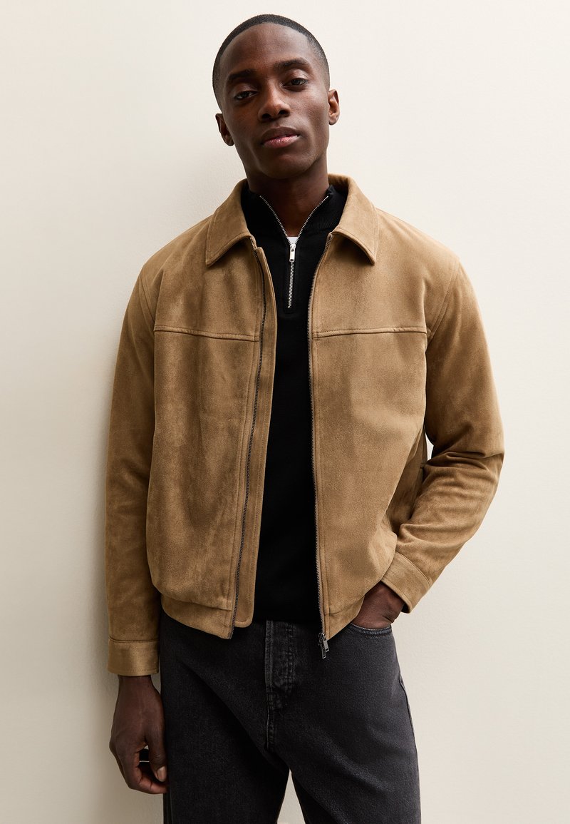 Uomo con giacca in camoscio color cognac sopra una camicia nera con zip, in piedi contro uno sfondo chiaro semplice con una mano in tasca.