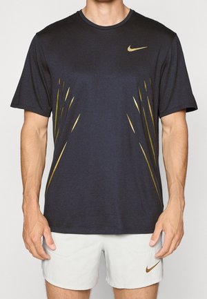 Homme portant un t-shirt Nike noir avec des accents dorés et un short blanc avec le logo doré Nike, debout devant un fond neutre.
