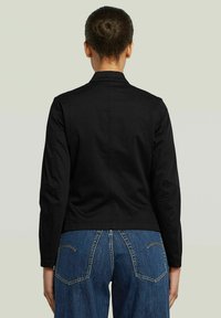 Giacca nera fitted con colletto alto, maniche lunghe e una texture liscia. Indossata sopra jeans in denim blu con dettagli di cucitura a contrasto.