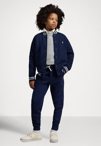 Marinblå fleece varsityjacka med vita randiga muddar och krage; grå sweatshirt under; marinblå sweatpants; cremevita sneakers; avslappnad passform.