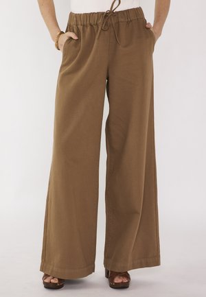 Pantaloni marroni a gamba larga con vita elasticizzata e coulisse, indossati con sandali, mani nelle tasche e una maglietta bianca visibile in vita.