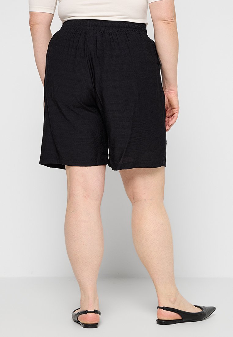 Schwarze Shorts mit elastischem Bund und rippigem Textur, die bis zur Mitte des Oberschenkels fallen, kombiniert mit schwarzen flachen Sandalen. Die Rückansicht zeigt Details der Beine.