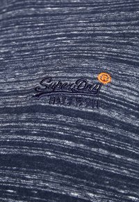 Námořnická modrá tkanina s texturovaným, melírovaným vzorem. Obsahuje vyšívané logo "SuperDry" ve tmavě fialové barvě, s oranžovým akcentem vedle něj.