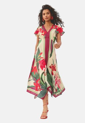 Sieviete pastaigājas krema krāsas kleitā ar lielu sarkanu un zaļu ziedu rakstu, V veida izgriezumu, plūstošām īsām piedurknēm un rozā siksniņu papēžiem.