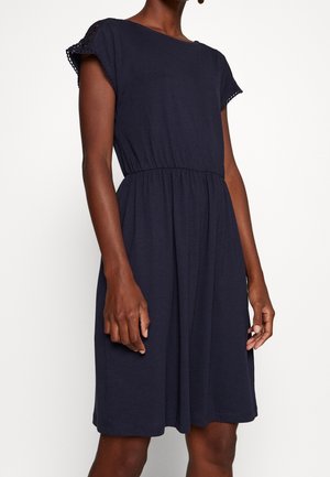 Femme portant une robe bleu marine mi-longue avec des manches courtes bordées de dentelle et une taille élastique froncée sur fond blanc.