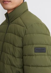 Olive green puffer jacket with a high collar, featuring quilted detailing and a black leather brand patch on the left sleeve.