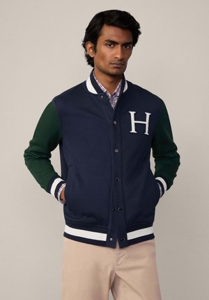 Marine varsityjack met groene mouwen, witte gestreepte zoom, drukknoopsluiting en een "H"-logo op de borst. Draag over een geruit shirt.