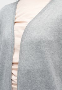 Ανοιχτό ροζ πουκάμισο που καλύπτεται εν μέρει από ένα ανοιχτό γκρι κάρντιγκαν, με έμφαση στην περιοχή του πάνω κορμού.