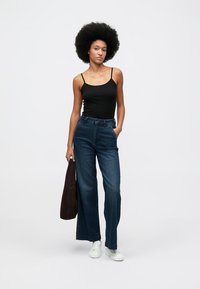 Svart tettsittende tanktopp, høy midje blå denim vide bukser, hvite joggesko og en mørk brun tote bag. Glatt, uformell design.