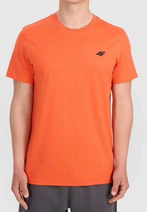Mężczyzna noszący jednolitą, jaskrawą pomarańczową koszulkę z krótkim rękawem z małym czarnym logo "4F" na lewej piersi, połączoną z ciemnymi spodniami.
