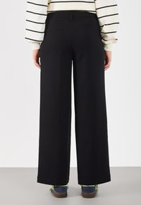 Melni plati bikses ar gludu tekstūru, ar augstu jostasvietu un divām aizmugures kabatām. Saskaņotas ar zilām kurpēm ar zaļiem akcentiem.