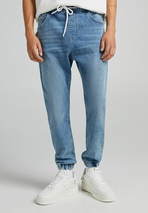 Man draagt lichtblauwe spijkerbroek jogger met elastische boorden, een wit trekkoord, witte sneakers en een wit overhemd.