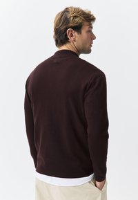 Mørkebrun strikket cardigan med rund krave, ribstrikkede ærmegab og en lige kant. Stoffet virker blødt og tekstureret.