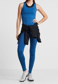 Modrý sportovní top bez rukávů a legíny s texturovanými akcenty, doplněné černým svetrem uvázaným v pase. Šedé tenisky dokončují vzhled.