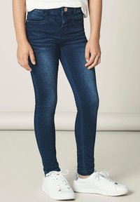 Jeans ajustados en azul oscuro con una cintura alta, fabricados en denim, que cuentan con dos bolsillos delanteros y una sutil textura desgastada, combinados con zapatillas blancas.