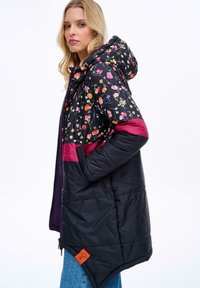 Zateplený kabát s květinovým vzorem v horní části, černou spodní částí, burgundskými akcenty a oranžovou nášivkou na lemu.