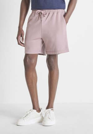 Uomo che indossa pantaloncini lilla chiaro con coulisse e sneaker bianche con lacci, in piedi davanti a uno sfondo chiaro e uniforme.