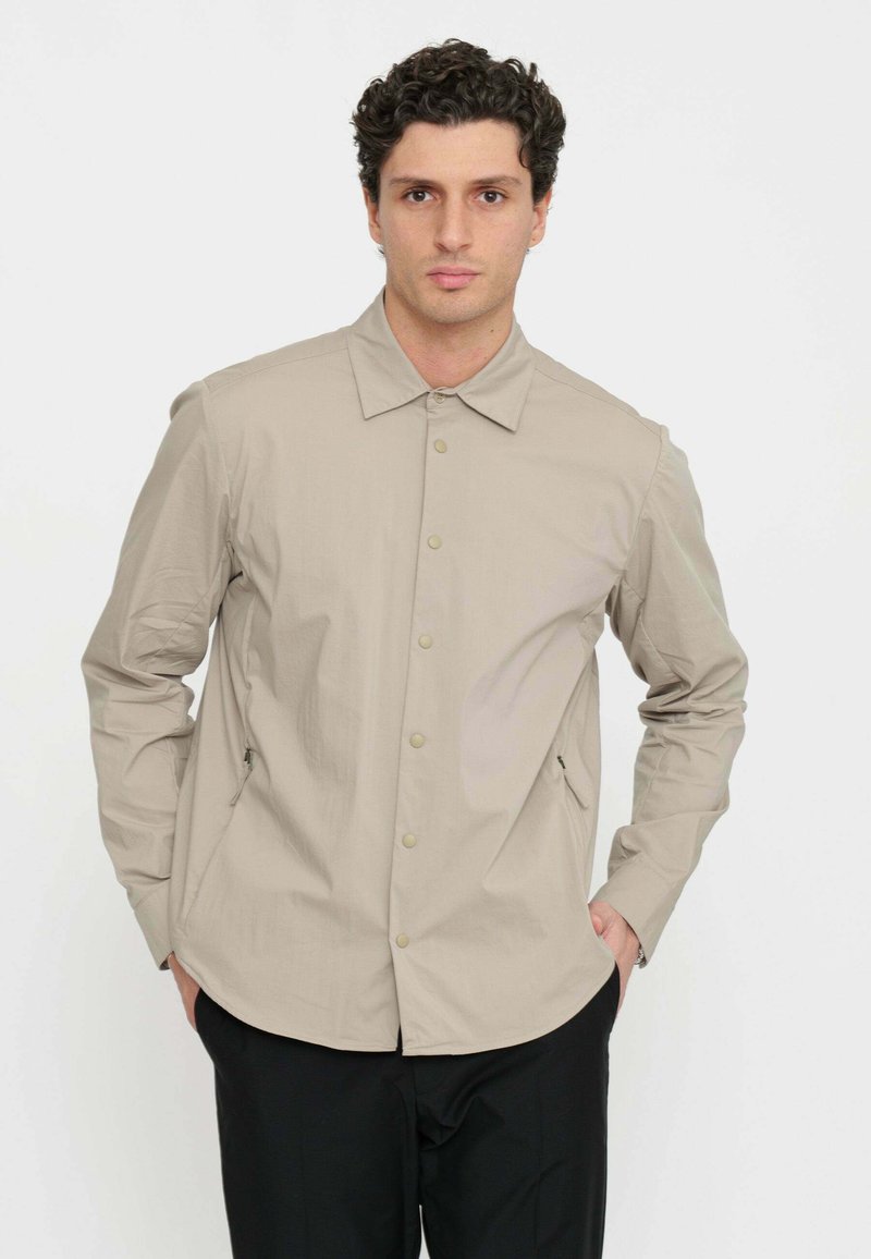 Camicia beige a maniche lunghe con apertura a bottoni, colletto a punta, tasche laterali e texture liscia. Indossata sopra pantaloni neri.
