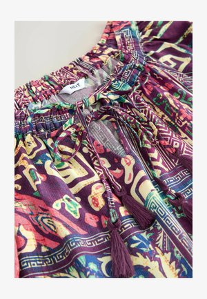 Blouse à motifs colorés avec une base violette, un col smocké, des liens et des accents à pompons ornés de motifs géométriques dans différentes teintes.