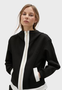 Crna dukserica s kapuljačom, bijelih patent zatvaračem i manžetama. Sadrži bočne džepove i izvezeni logo detalj na prsima. Glatka tekstura.