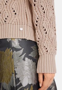 Strickpullover in hellem Beige mit texturiertem Muster und gerippten Bündchen, kombiniert mit einem dunklen, floralen Stoffmuster in Silber- und Grüntönen.