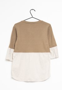 Camicia a maniche lunghe con parte superiore in un solido colore beige e parte inferiore a righe bianche. Materiale in cotone, orlo arrotondato e vestibilità comoda. Appesa a un gancio nero.