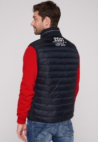 Gilet imbottito blu navy con cuciture orizzontali, testo bianco sul retro e collo alto. Indossato sopra una maglietta rossa a maniche lunghe.