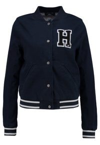 Námorně modrá bomber bunda vyrobená z odolné látky, s bílou nášivkou "H", sponami na druky a pruhovanými manžetami a lemem.