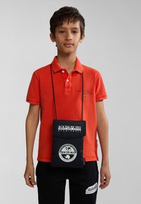 Polo arancione con colletto, abbinato a pantaloni neri con un'etichetta logo. Una borsa a tracolla nera in tessuto pende da un cordino.