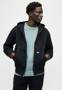 Νέος άνδρας που κοιτάζει πλάγια, φοράει μαύρο φούτερ με φερμουάρ πάνω από ανοιχτό μπλε πουλόβερ, τα χέρια στις τσέπες, μπροστά σε απλό γκρι φόντο.