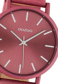 Pink watch with a round metal case, textured strap, minimalist silver hour markers, and logo at 12 o'clock. Smooth dial finish.