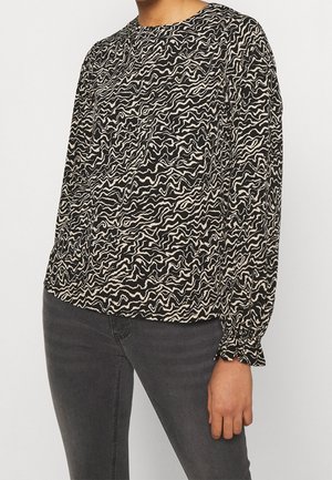 Femme portant un chemisier à manches longues avec un motif abstrait noir et blanc et des poignets volantés, associé à un jean gris foncé.