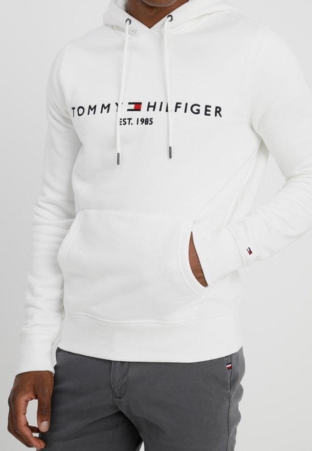 Sweat à capuche blanc Tommy Hilfiger avec logo et drapeau sur la poitrine, main dans la poche avant, associé à un pantalon gris foncé.
