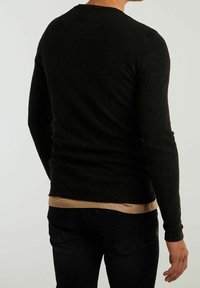 Ciemnozielona, fakturowana koszula z długim rękawem, z okrągłym dekoltem, noszona na beżowej koszuli, w zestawieniu z czarnymi dżinsami, widok z tyłu.