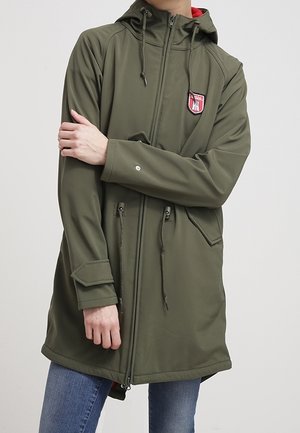 Personne portant une veste à capuche vert olive avec un écusson, une fermeture éclair, des cordons de serrage, des poignets à rabat, et un jean bleu, les bras croisés sur la poitrine.