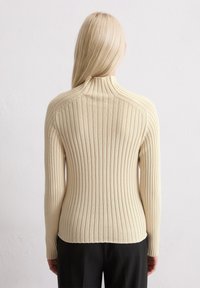 Beige geribde trui met een hoge hals, voorzien van lange mouwen en een aansluitende silhoutte. De stof heeft een gladde textuur met verticale lijnen.
